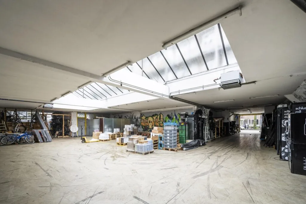 Ruime industriële opslagruimte aan de Baarsjesweg met skylights, graffiti op de muur en gestapelde materialen op pallets.