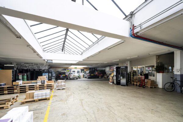 Magazijnruimte aan de Baarsjesweg met geparkeerde auto's, stapels dozen op pallets en een glazen dak voor natuurlijk licht.