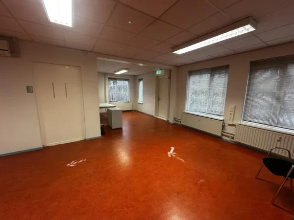 Een leegstaand kantoor met roodbruine vloer, witte muren, bureaus bij het raam en enkele stoelen, in gebouw Veulenkamp.