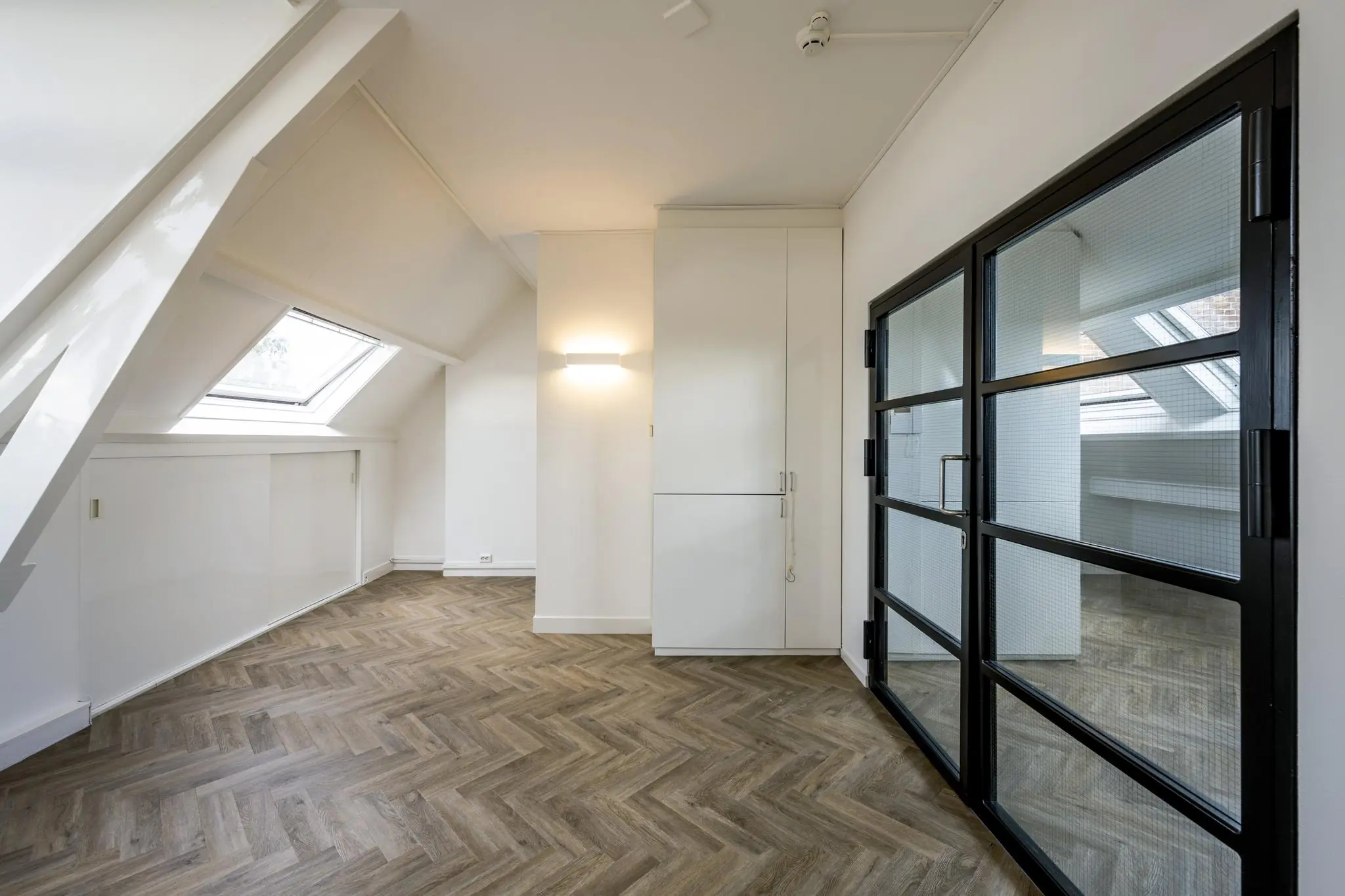 Lichte zolderruimte op het Emmaplein met visgraatvloer, schuine dakramen en zwarte glazen deuren.
