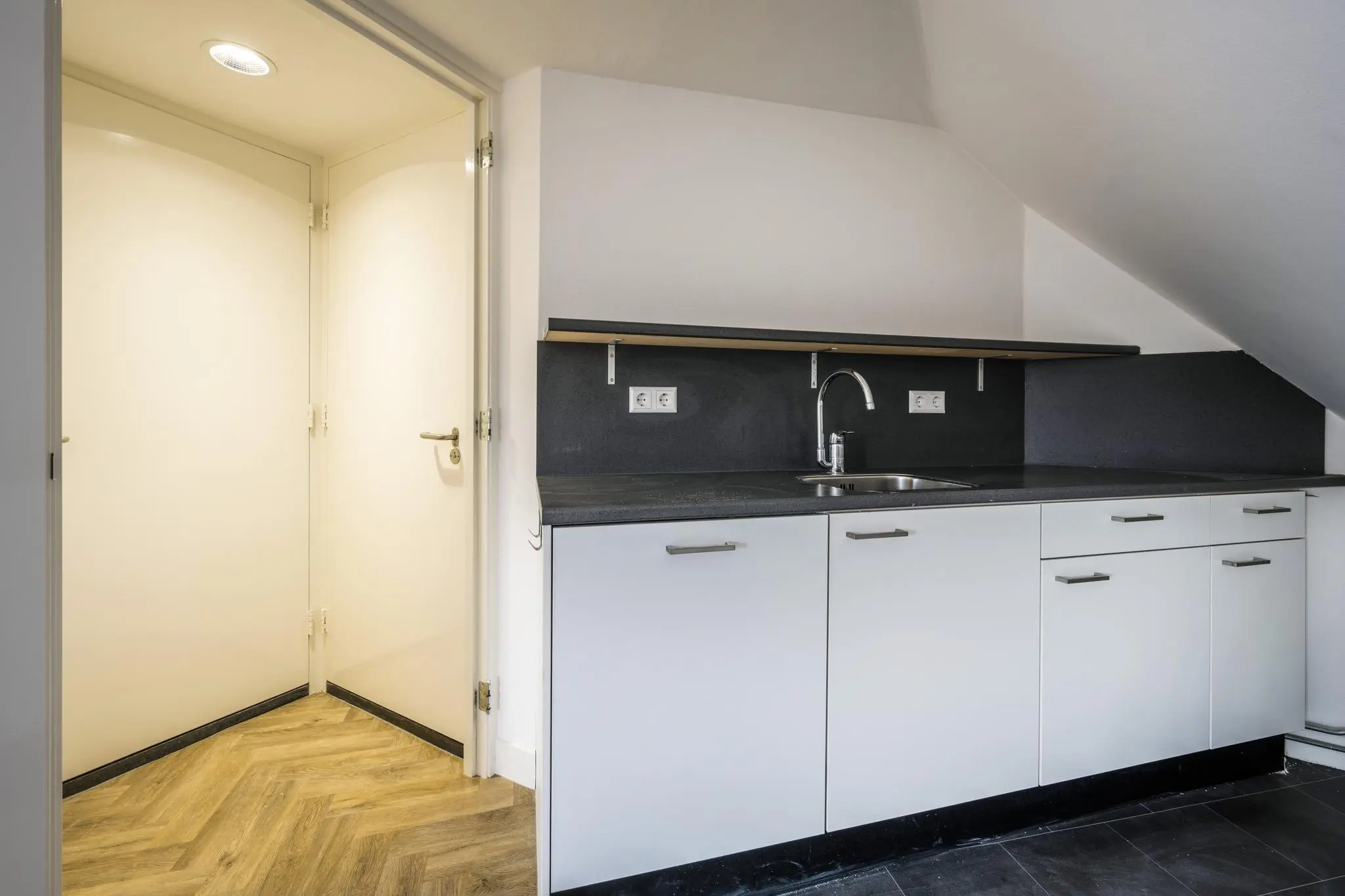 Witte kitchenette onder een schuine wand met zwart werkblad en gootsteen, naast een open deur naar een hal met houten visgraatvloer.