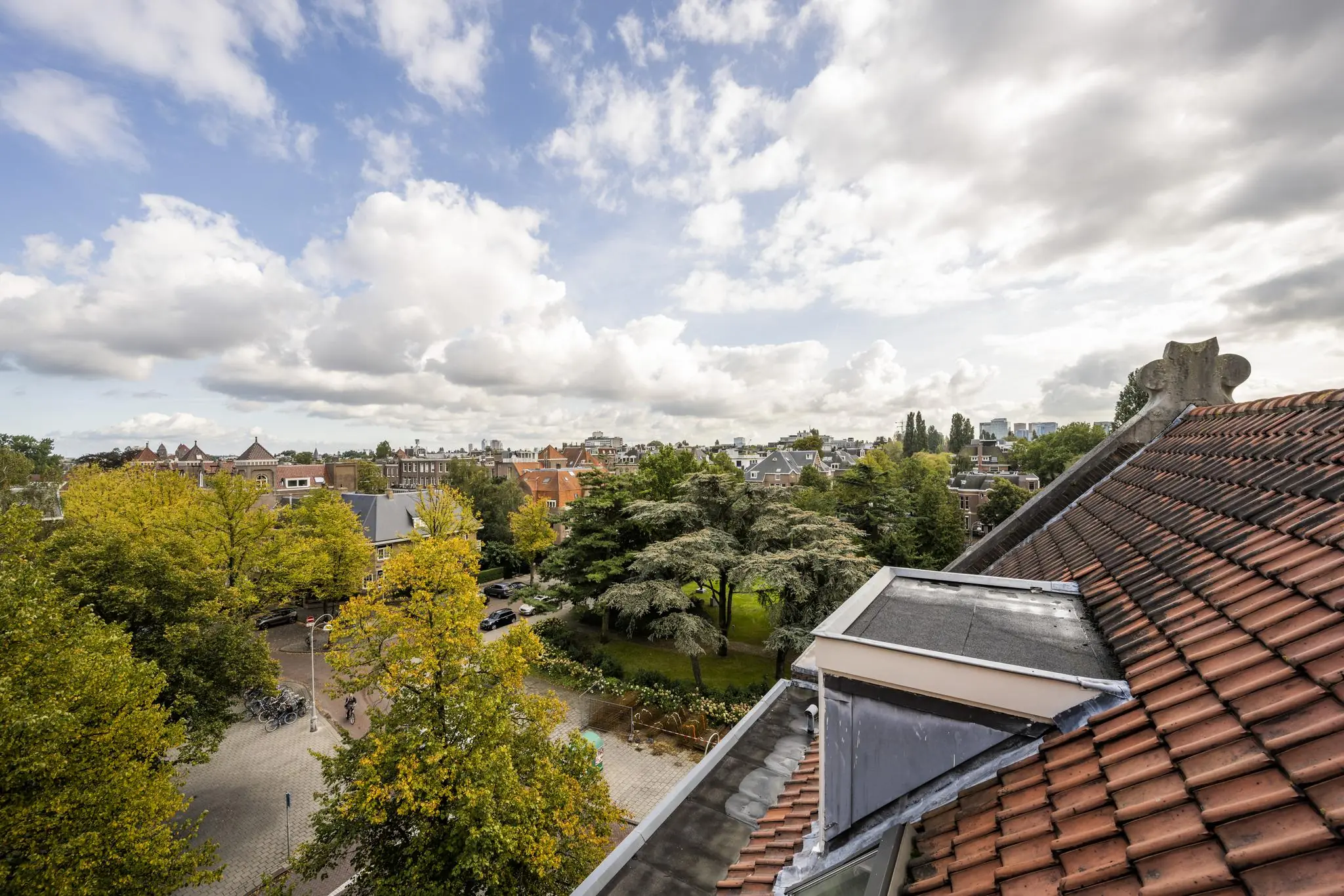 Uitzicht op het Emmaplein in Amsterdam met groene bomen, historische gebouwen en een pannendak op de voorgrond onder een deels bewolkte hemel.