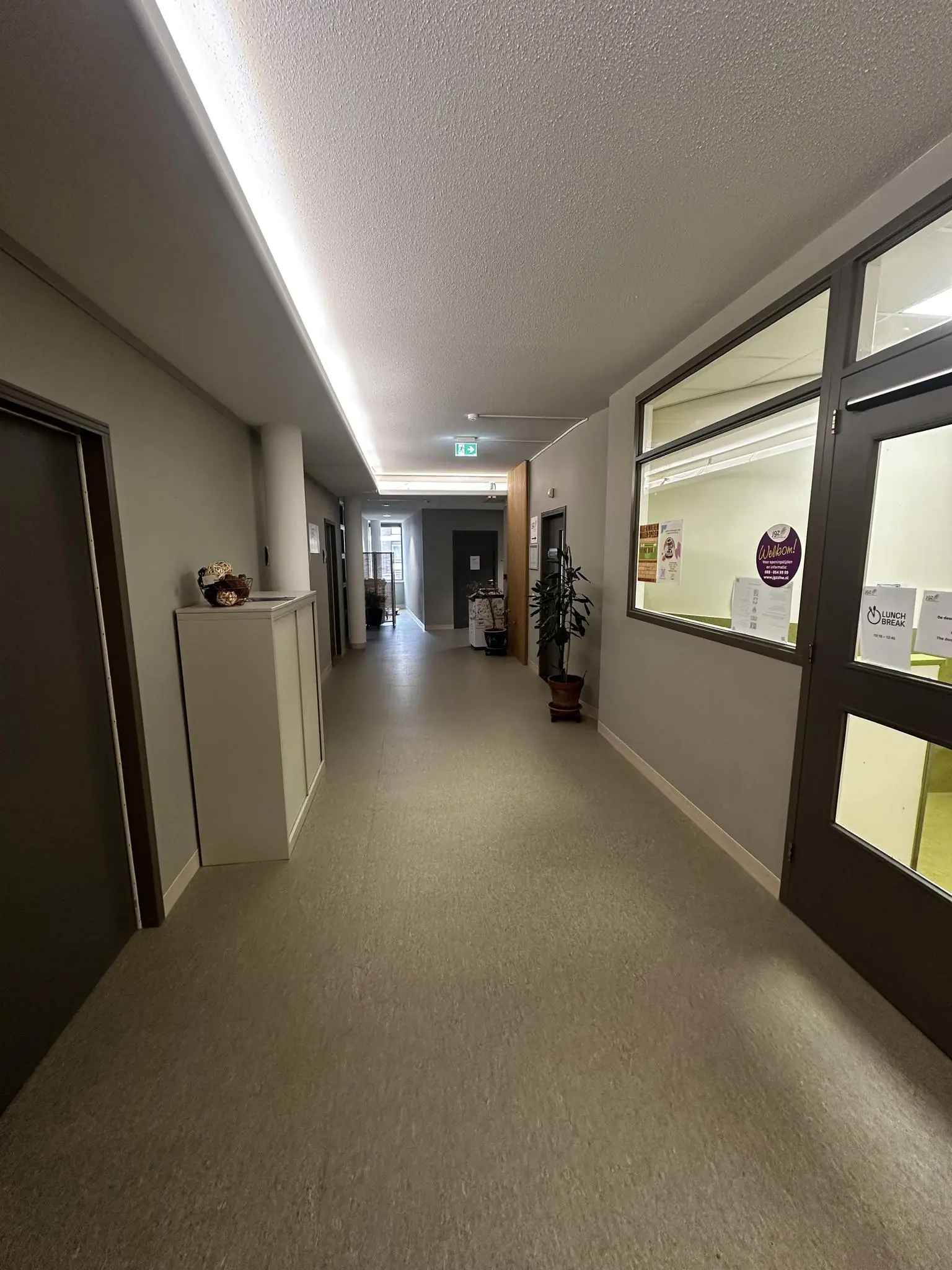 Verlichte gang in een gebouw aan de Nassaulaan met deuren, planten en informatieve posters aan de muur.