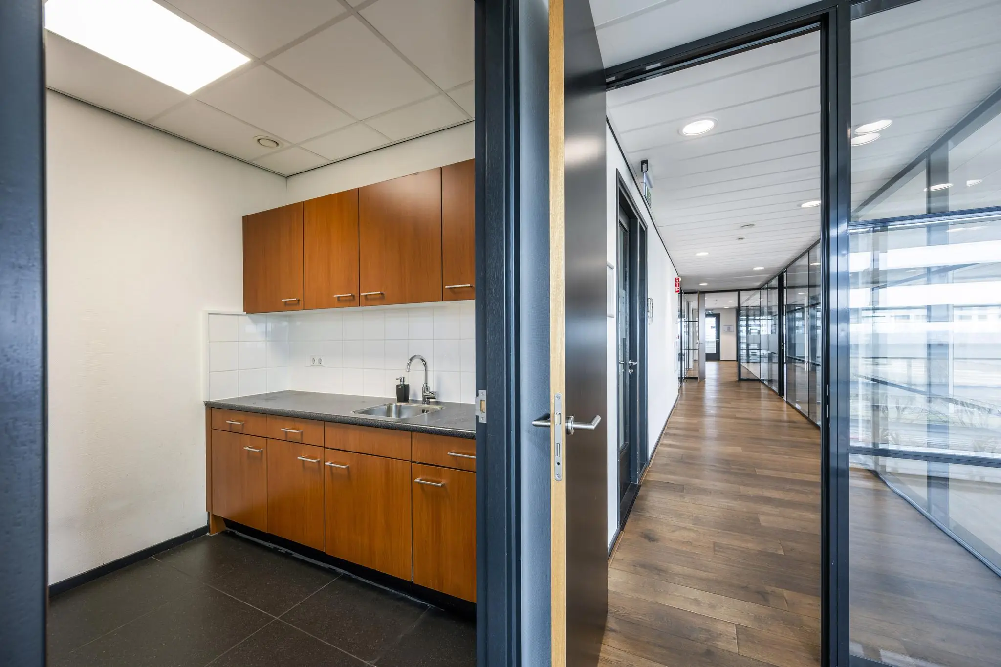Kleine kantoorpantry met houten keukenkastjes en uitzicht op een moderne gang met glazen wanden in het Sijsjesbergweg-kantoor.