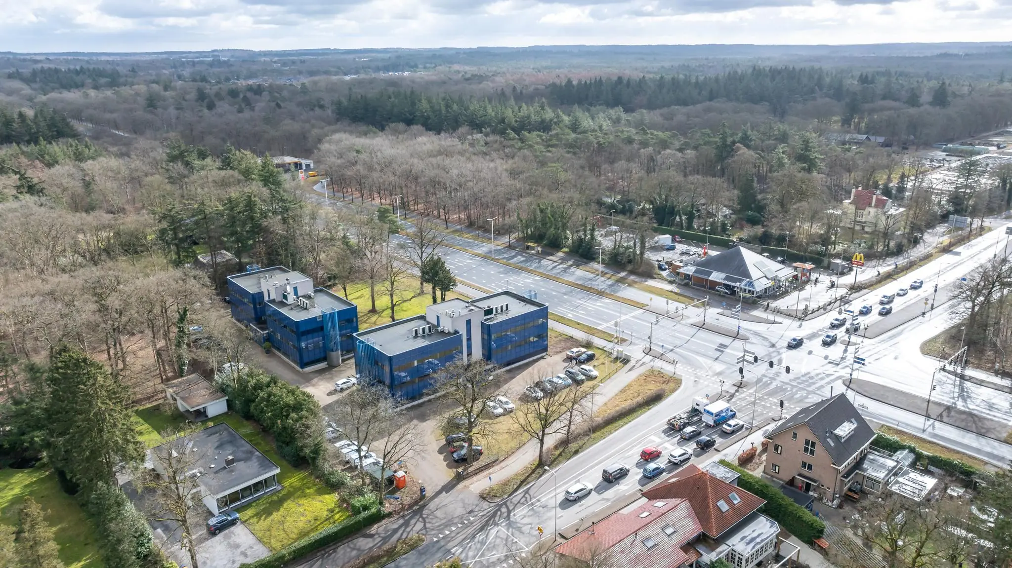 Luchtfoto van de Dolderseweg met blauwe kantoorgebouwen, een kruispunt en een McDonald's, omringd door bosgebied.