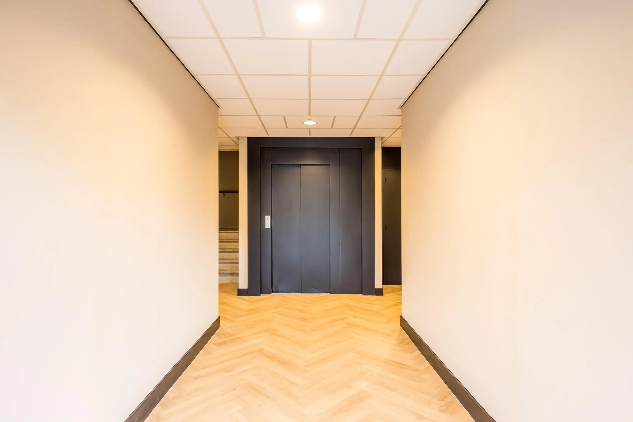 Verlichte gang met houten visgraatvloer en een zwarte liftdeur aan het einde, locatie Dolderseweg.