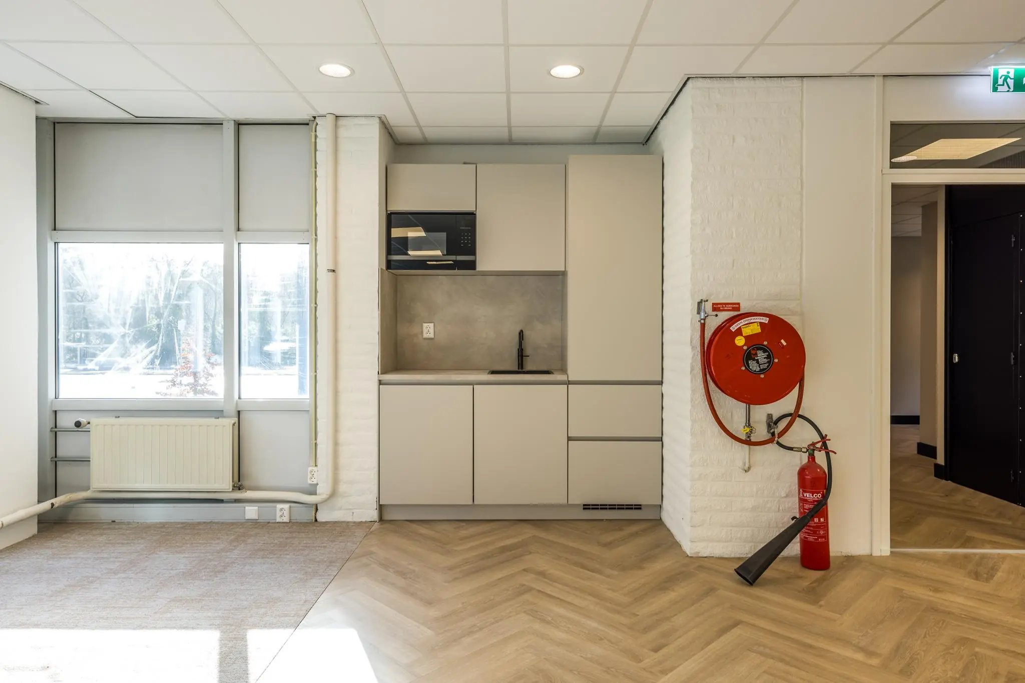 Moderne pantry met lichtgrijze keukenkasten, een ingebouwde magnetron en een brandslanghaspel aan de muur in een kantoorpand aan de Dolderseweg.