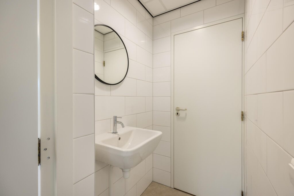 Wit betegelde toiletruimte met een wandfontein, ronde spiegel en witte deur.