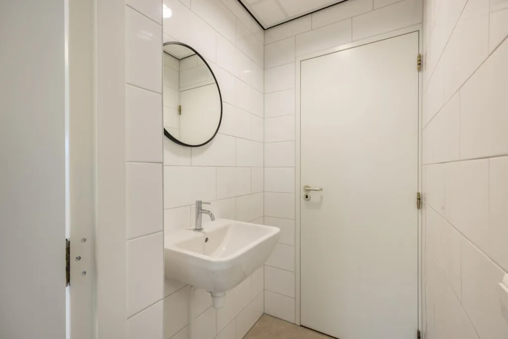 Wit betegelde toiletruimte met een wandfontein, ronde spiegel en witte deur.