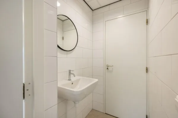 Wit betegelde toiletruimte met een wandfontein, ronde spiegel en witte deur.