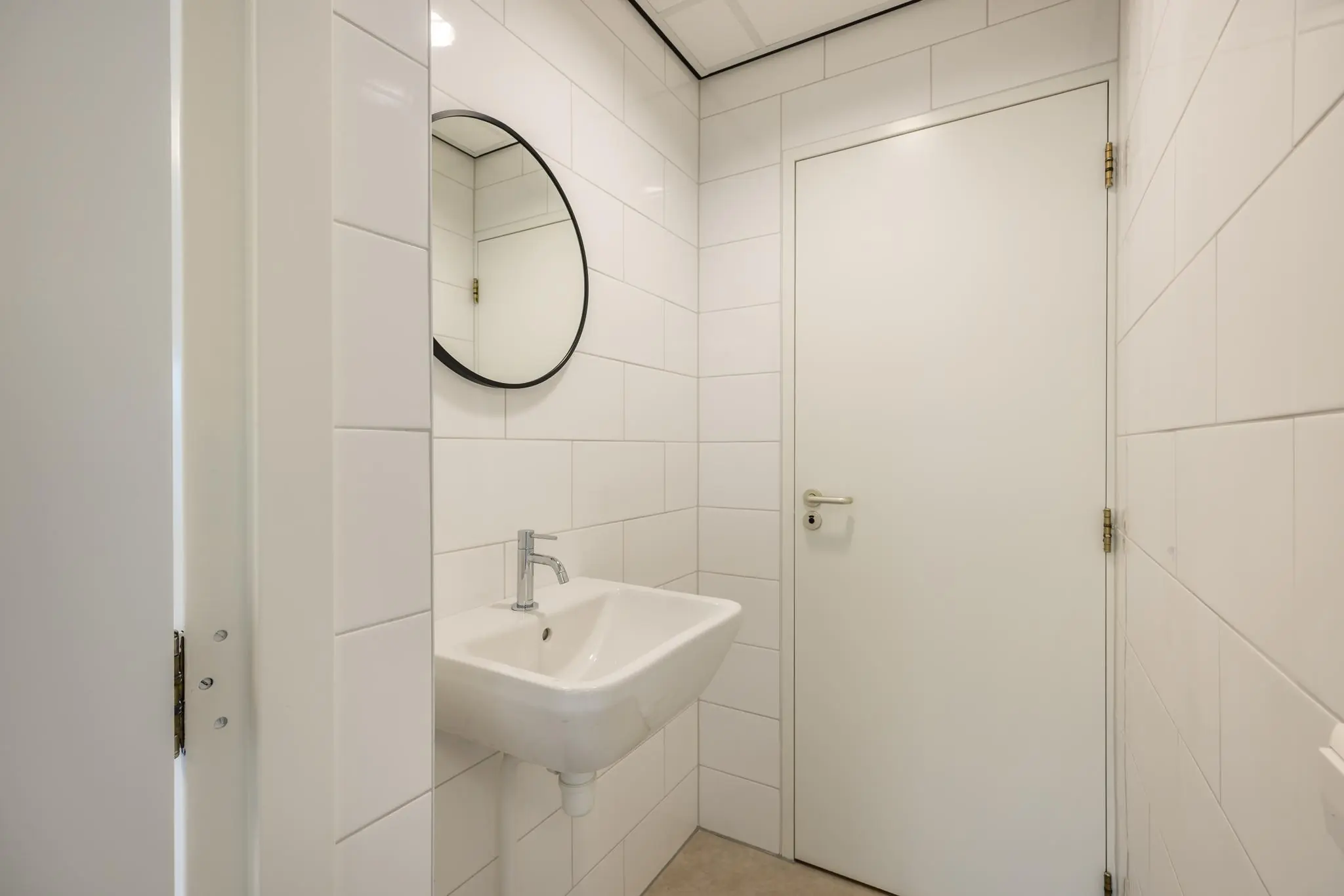 Wit betegelde toiletruimte met een wandfontein, ronde spiegel en witte deur.