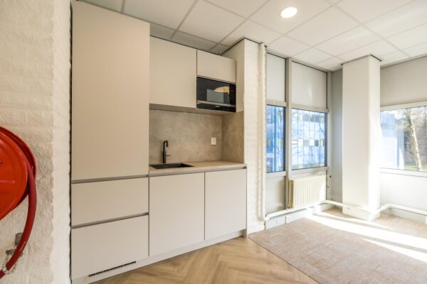 Moderne kitchenette met beige kasten, zwarte kraan en ingebouwde magnetron in een lichte kantoorruimte aan de Dolderseweg.