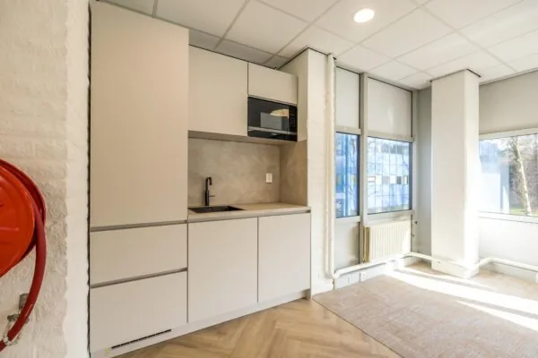 Moderne kitchenette met beige kasten, zwarte kraan en ingebouwde magnetron in een lichte kantoorruimte aan de Dolderseweg.