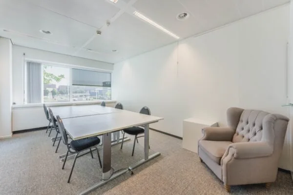 Vergaderruimte aan de Laarderhoogtweg met een tafel, zes stoelen, een grote fauteuil en een raam met uitzicht op een bedrijventerrein.