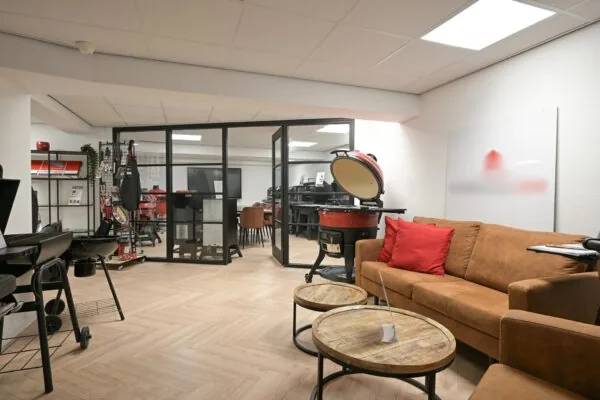 Showroom met barbecues en accessoires, ingericht met een bruine bank, houten tafels en een glazen vergaderruimte op de achtergrond.