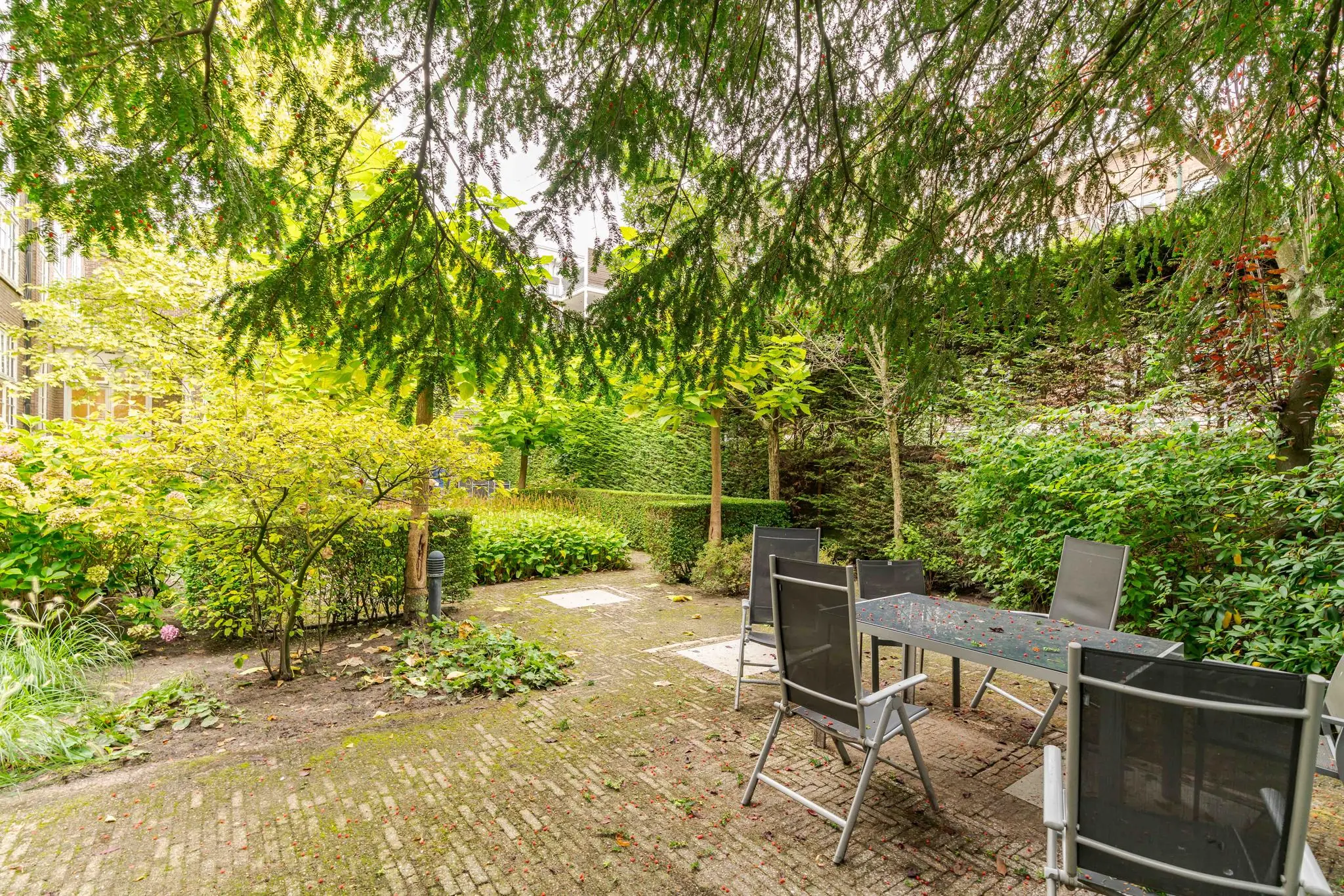 Binnentuin aan het Lange Voorhout met een betegeld terras, tuintafel en stoelen, omgeven door weelderig groen en hoge hagen.