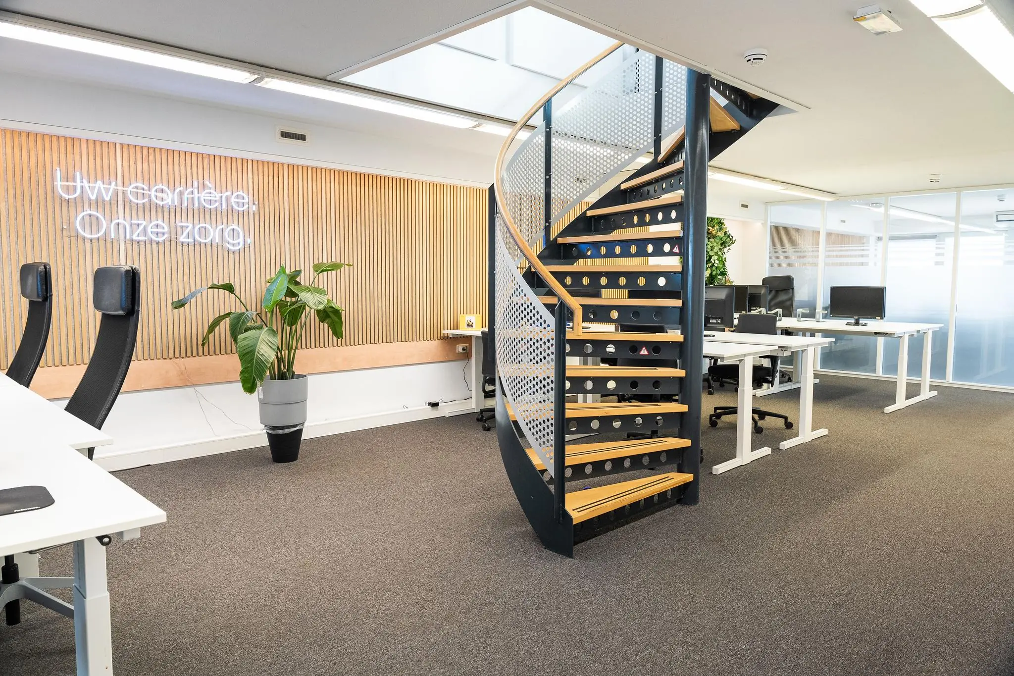 Modern kantoorinterieur aan de Prins Hendrikkade met een wenteltrap, bureaus en de tekst "Uw carrière. Onze zorg." aan de muur.