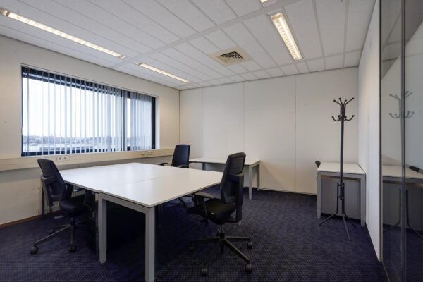 Kantoorruimte aan de Laan van ’s-Gravenmade met bureaus, bureaustoelen, een kapstok en een groot raam met verticale lamellen.