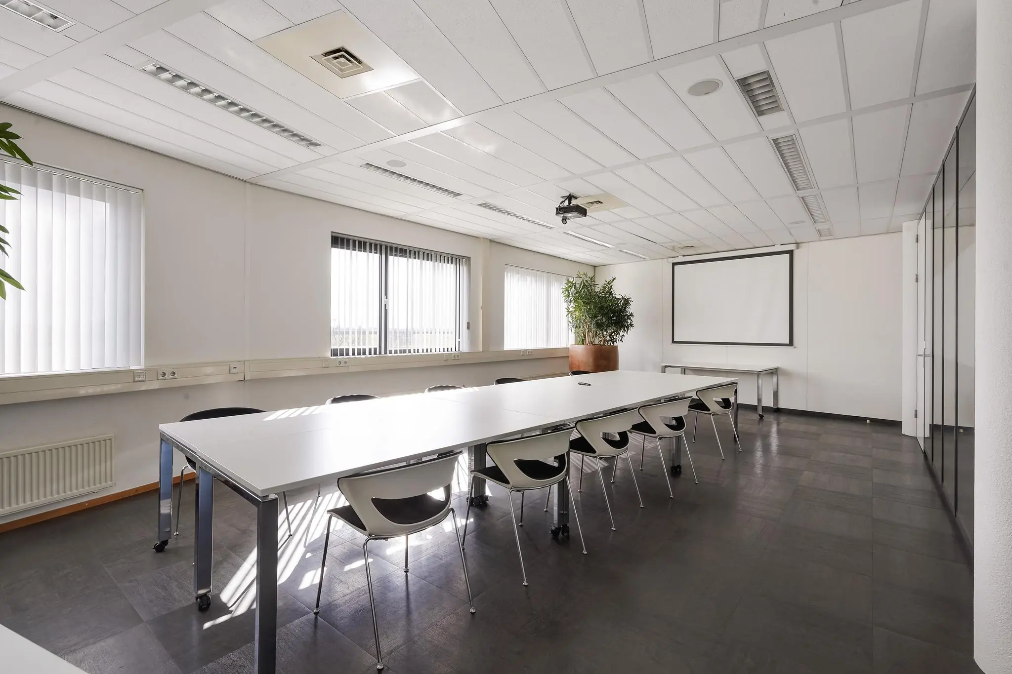 Vergaderruimte aan de Laan van S-Gravenmade met een lange tafel, moderne stoelen, een beamer en een projectiescherm.