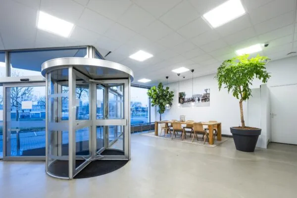 Moderne entreehal aan de Van der Madeweg met een glazen draaideur, zithoek met houten meubilair en grote kamerplanten.