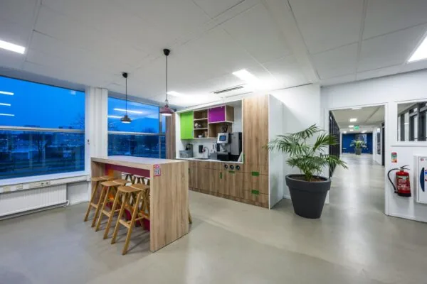 Moderne kantoorkeuken aan de Van der Madeweg met houten bar, barkrukken, koffieautomaat en grote plant in een lichte open ruimte.