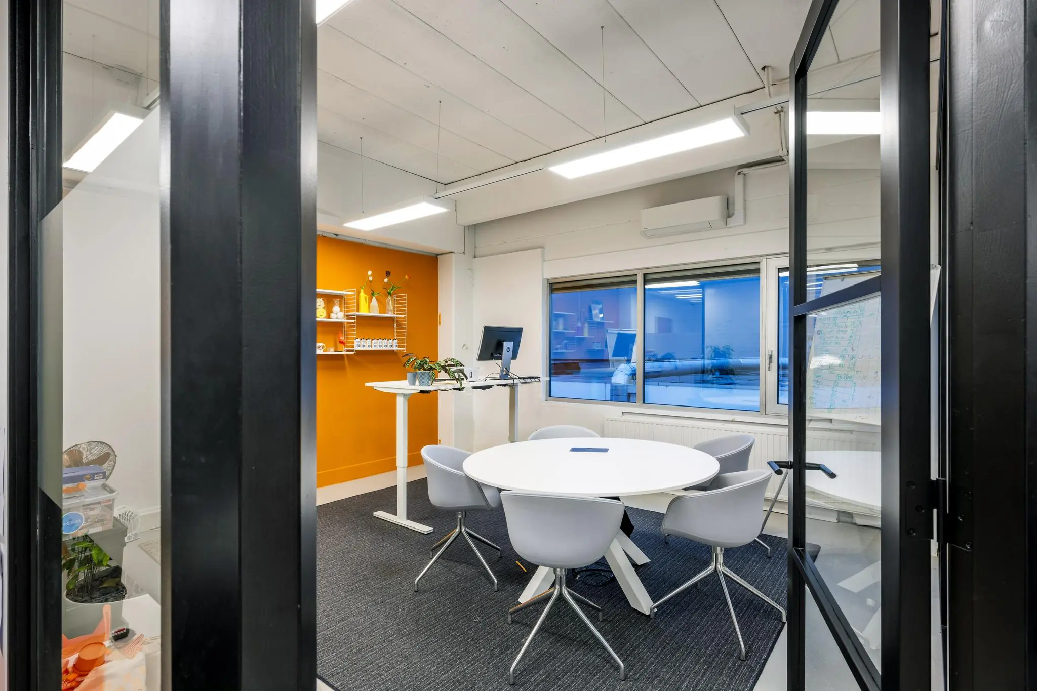 Moderne vergaderruimte aan de Van der Madeweg met ronde tafel, vijf stoelen, oranje accentmuur en verstelbaar bureau met monitor.