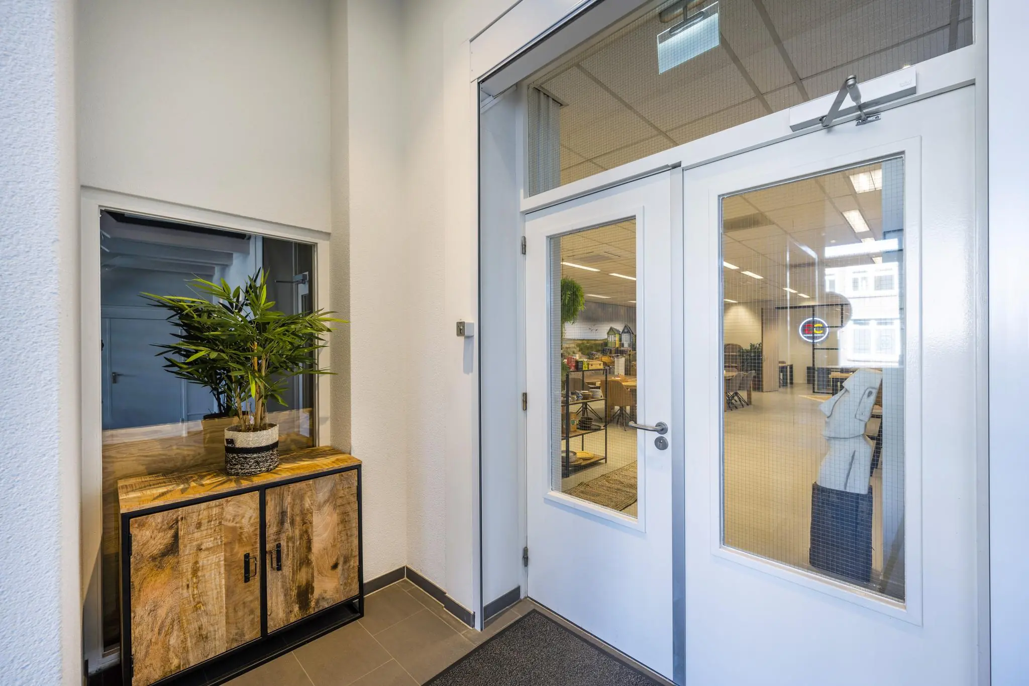 Ingang van een modern kantoorpand aan de Helicopterstraat met glazen dubbele deuren en houten kast met plant.