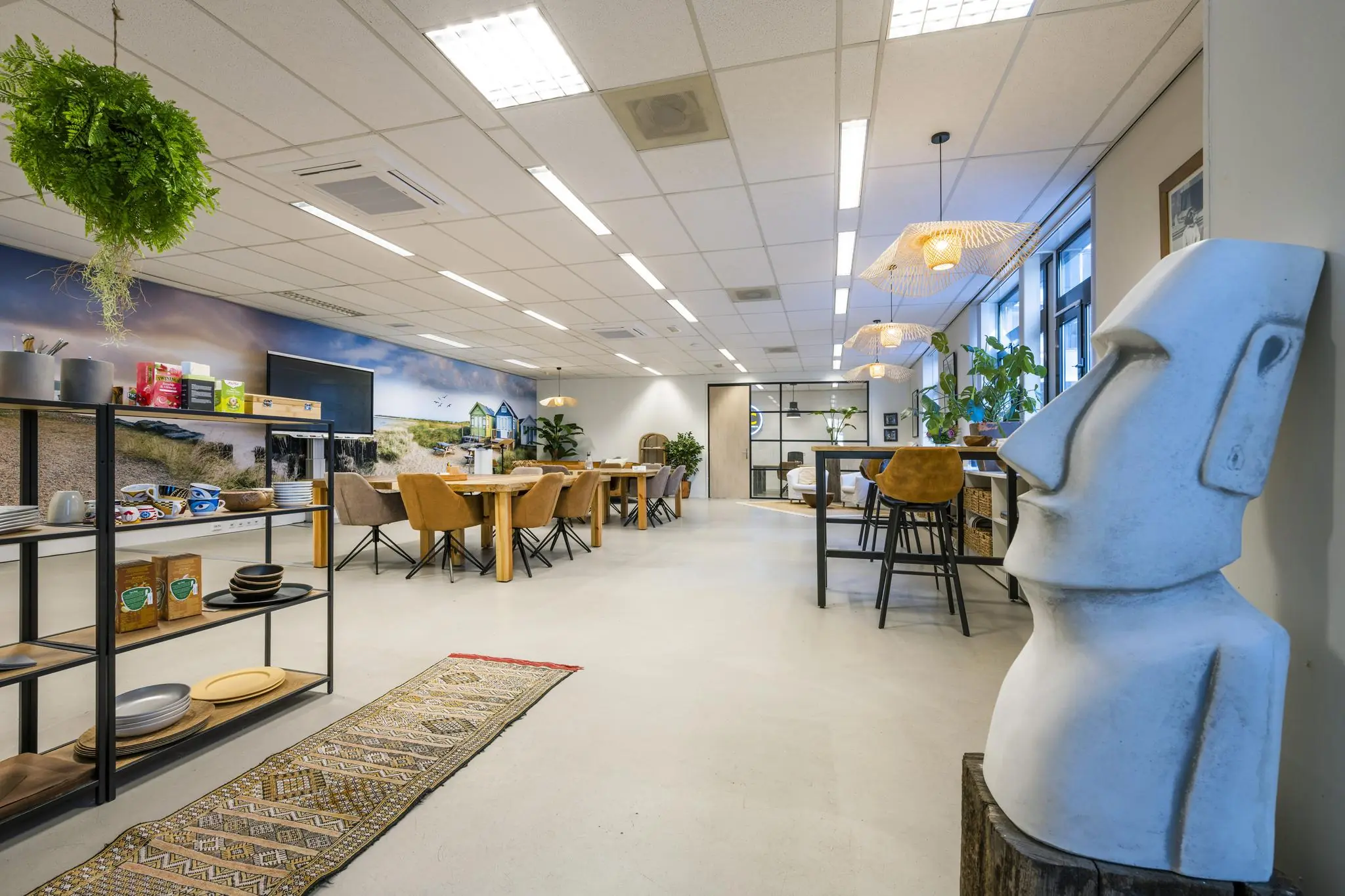 Modern ingerichte kantooruimte aan de Helicopterstraat met houten meubels, planten, een grote muurfoto van strandhuisjes en een opvallend Moai-beeld.