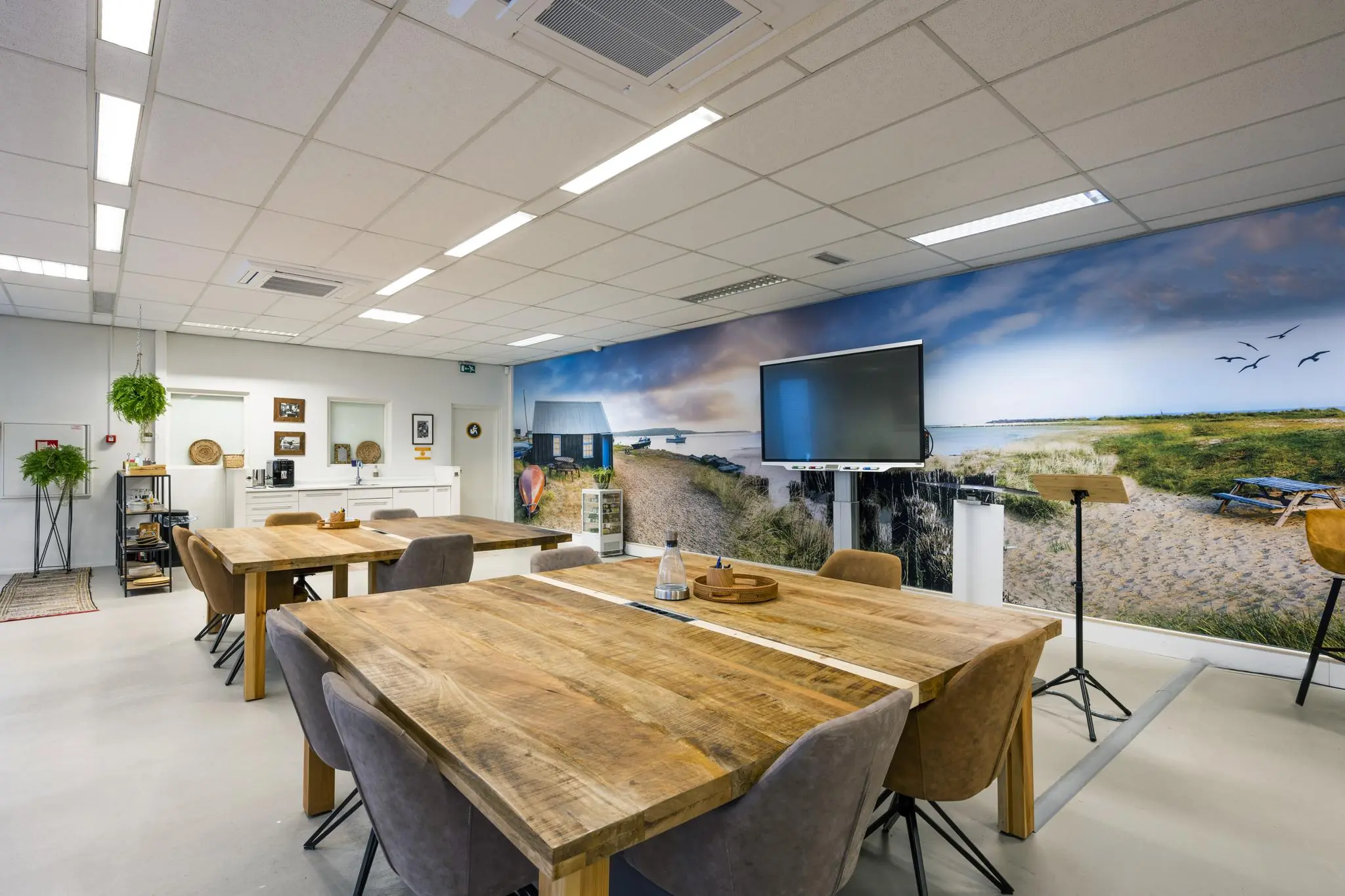 Moderne vergaderruimte met houten tafels, een groot scherm en een strandlandschap als muurdecoratie.