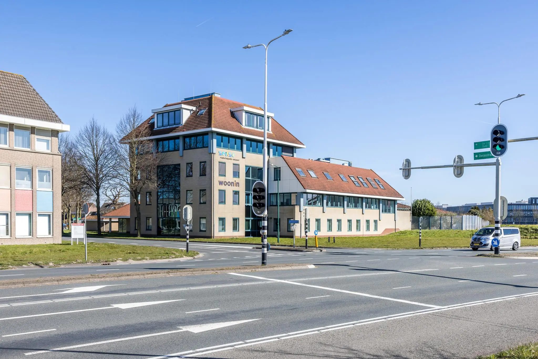 Kantoorgebouw van Woonin aan de Dorpsstraat op een zonnige dag, gezien vanaf een kruispunt met verkeerslichten.