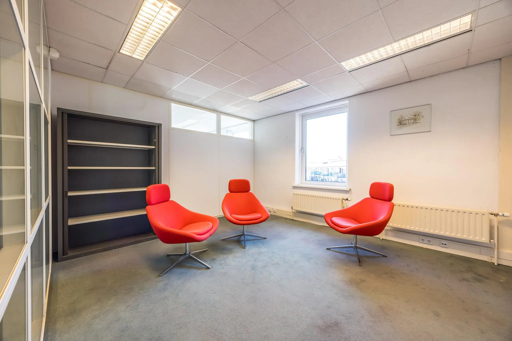 Vergaderruimte met drie moderne rode stoelen, een leeg zwart wandrek en een raam dat daglicht binnenlaat.