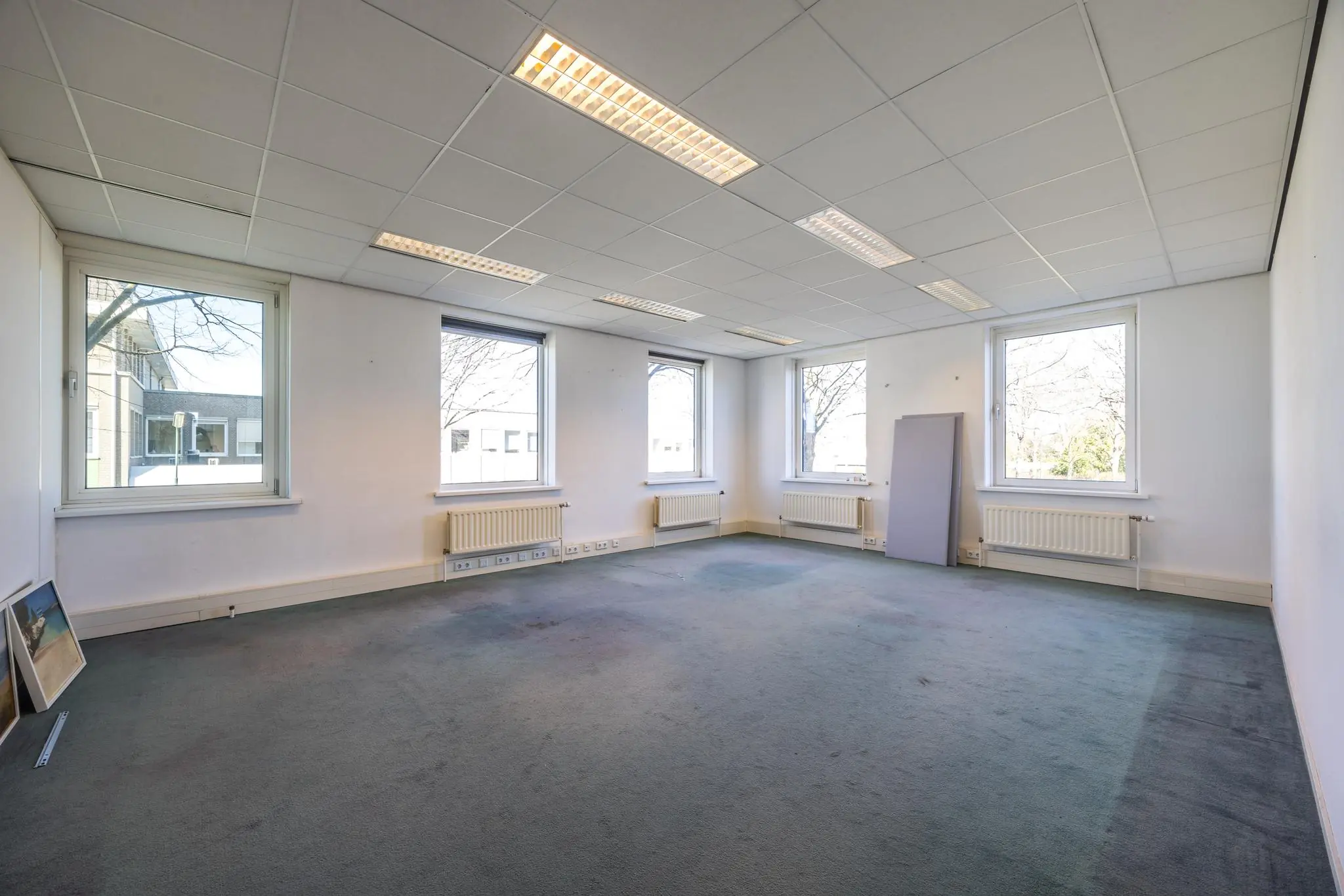 Lege kantoorruimte aan de Dorpsstraat met vier grote ramen, systeemplafond en blauw tapijt.