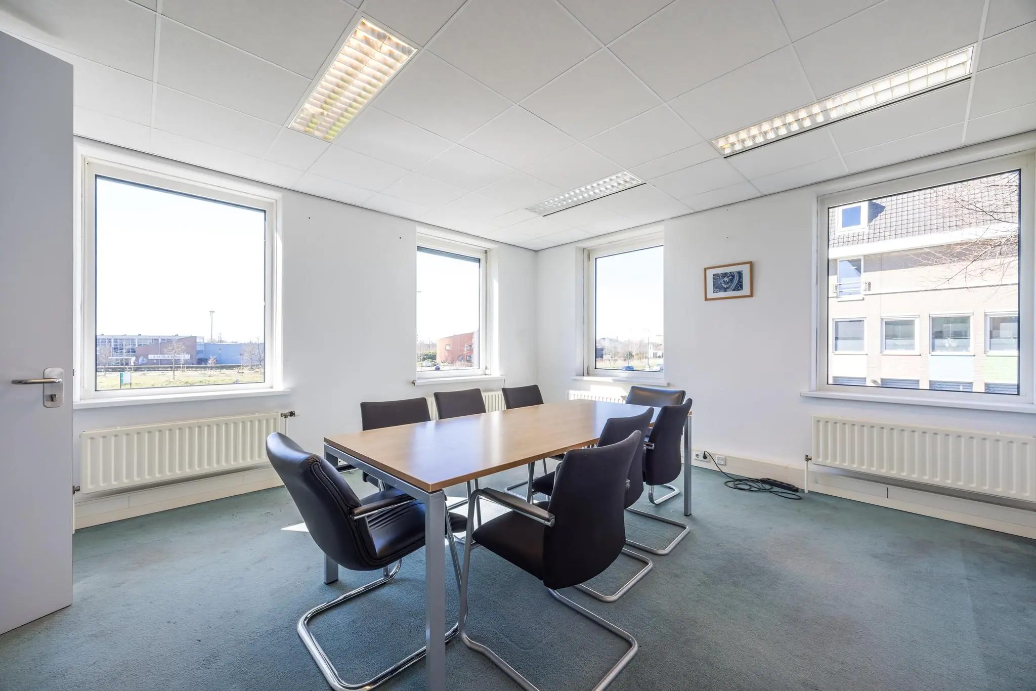 Vergaderruimte met zes stoelen rond een houten tafel in een kantoorpand aan de Dorpsstraat, met veel daglicht via vier grote ramen.
