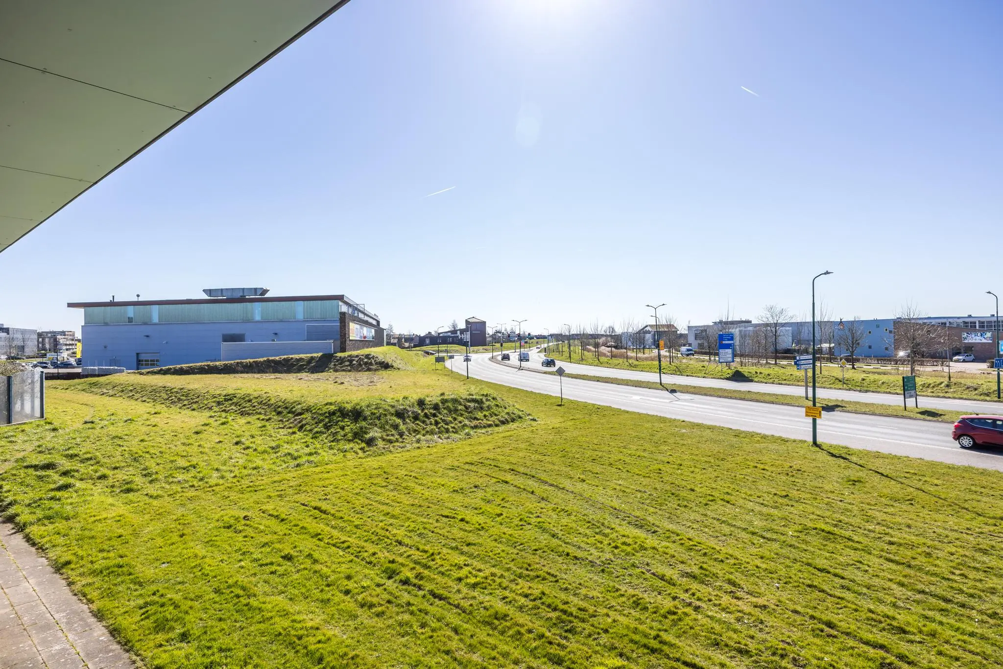 Moderne straatzicht op de Dorpsstraat met grasvelden, een blauw gebouw en enkele rijdende auto's onder een heldere lucht.