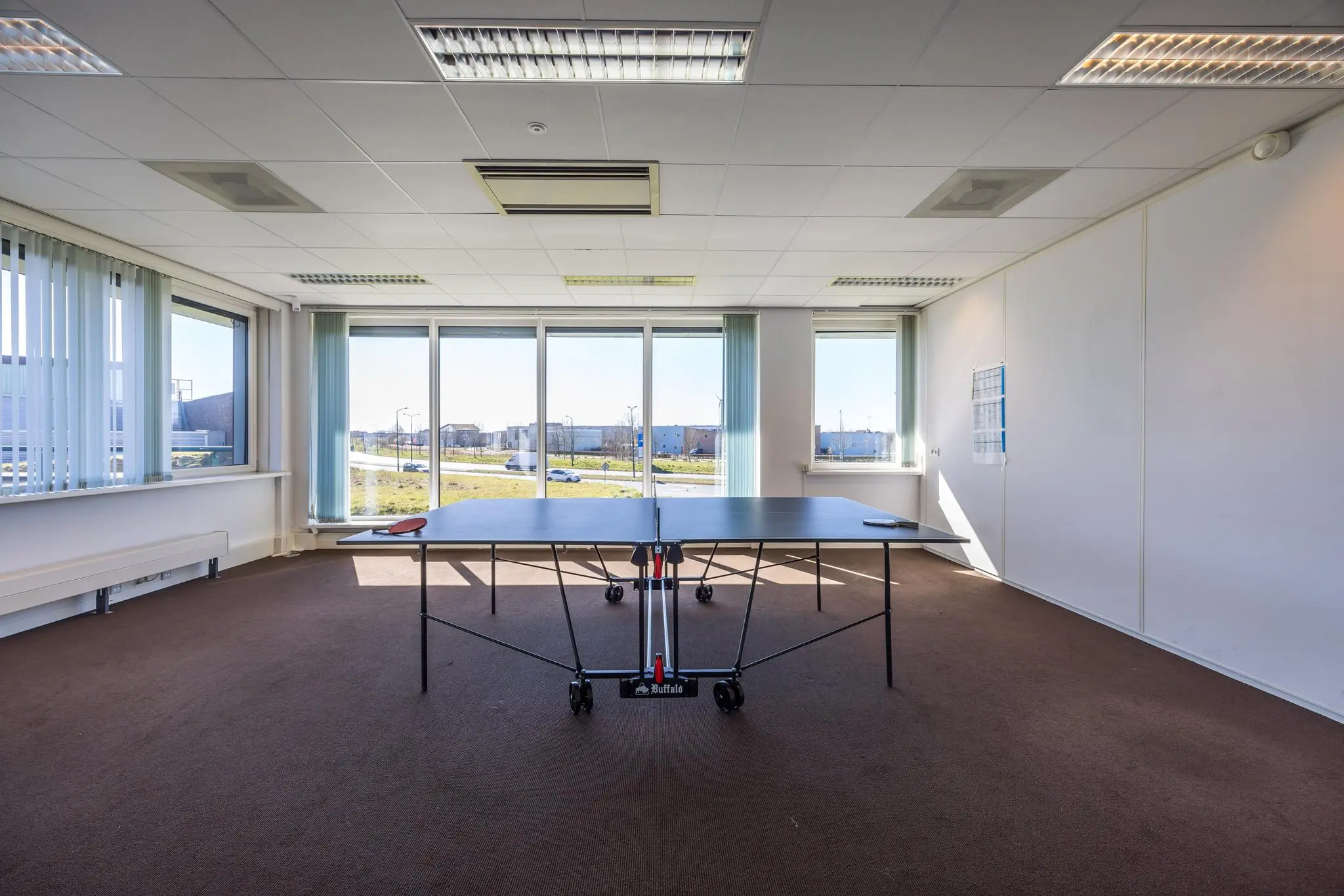 Kantoorruimte met een pingpongtafel in het midden en grote ramen met uitzicht op een bedrijventerrein aan de Dorpsstraat.