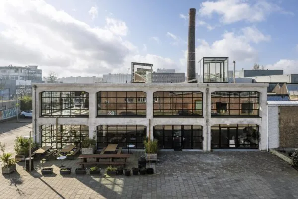 Vooraanzicht van een industrieel pand aan het Gedempt Hamerkanaal met grote ramen en een terras met houten picknicktafels.