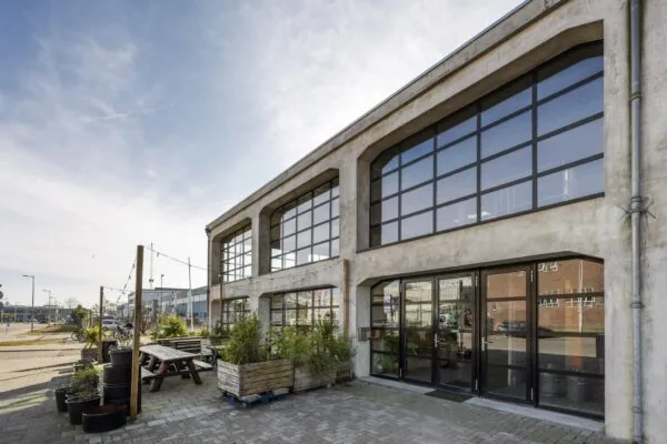 Industriële gevel van een gerenoveerd pand aan het Gedempt Hamerkanaal met grote ramen en een buitenzitgedeelte met planten.