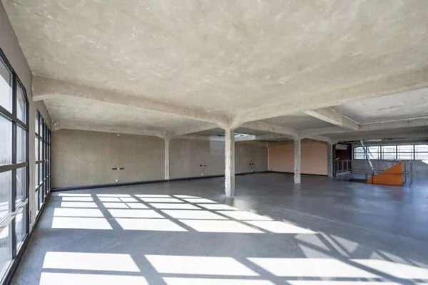 Lichte, lege industriële ruimte in het Gedempt Hamerkanaal met betonnen vloer en plafonds, grote ramen en een trap naar beneden.