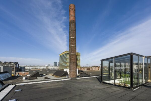 Uitzicht vanaf een plat dak aan het Gedempt Hamerkanaal met een bakstenen fabrieksschoorsteen, moderne gebouwen en een glazen trapopgang.