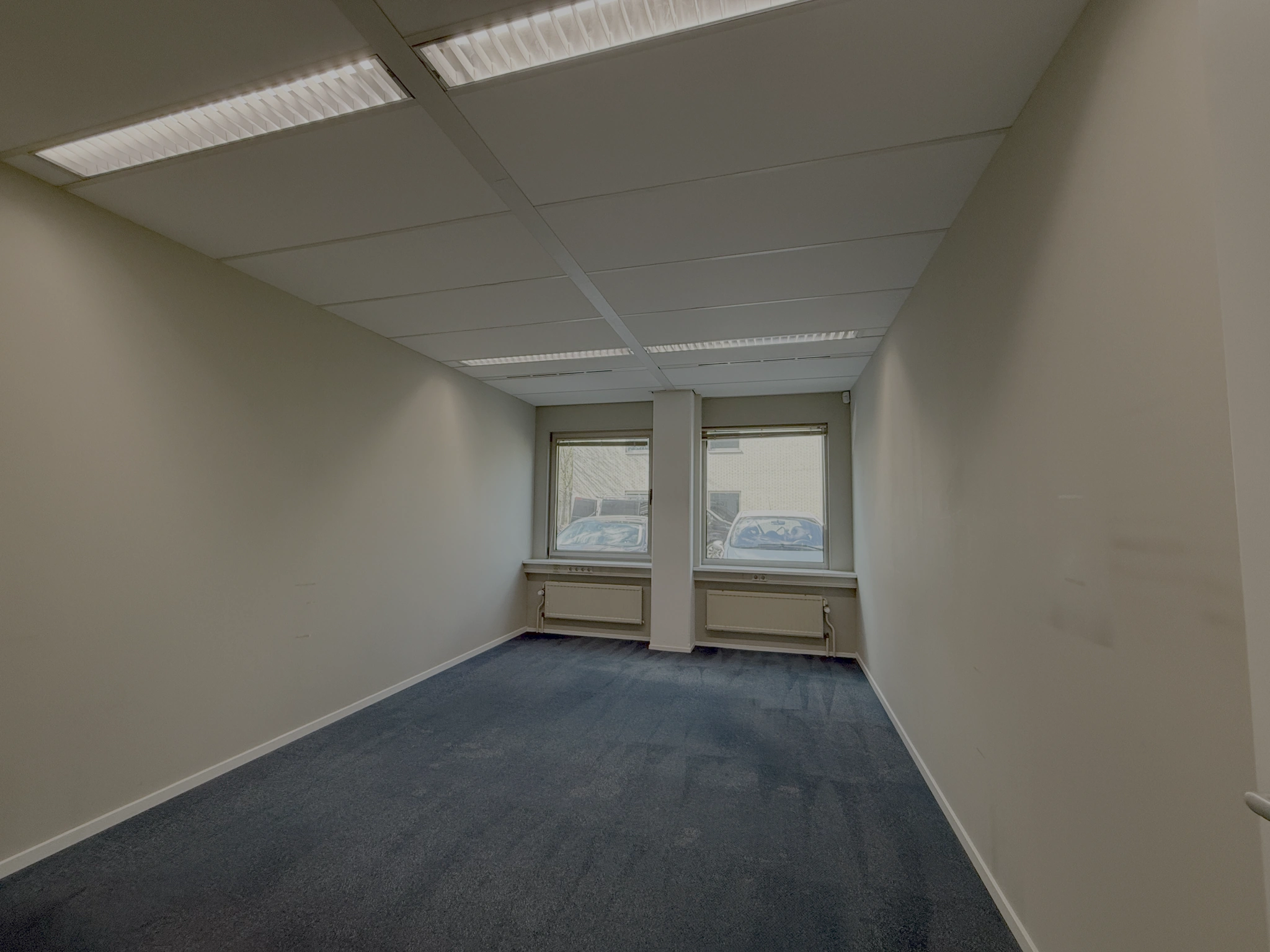 Lege kantoorruimte aan de Lunettenbaan met blauw tapijt, witte muren en twee ramen aan de straatzijde.