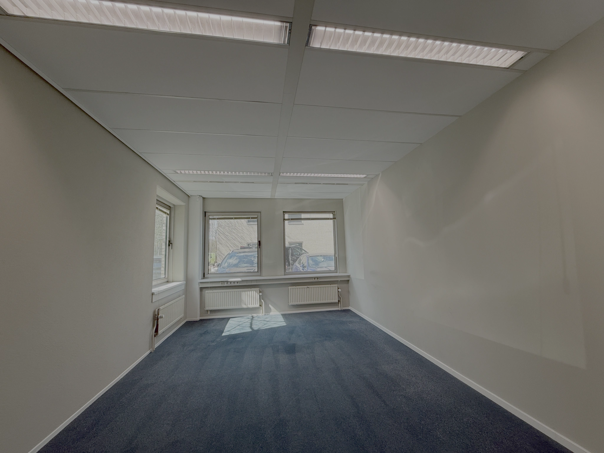 Lege kantoorruimte aan de Lunettenbaan met blauw tapijt, witte muren en drie ramen aan de voorzijde.