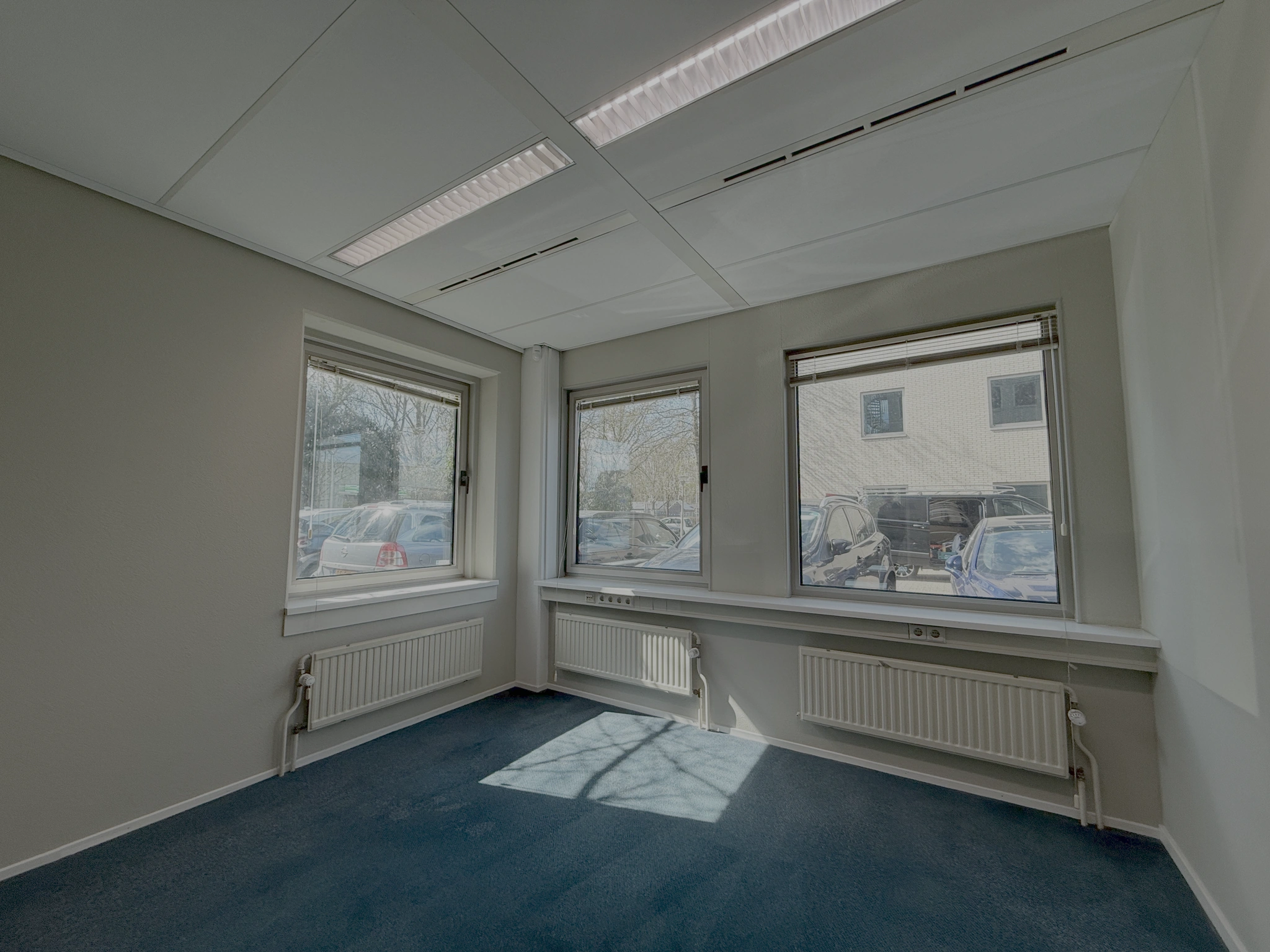 Leeg kantoor met drie ramen aan de Lunettenbaan, blauwe vloerbedekking en zicht op parkeerplaatsen buiten.