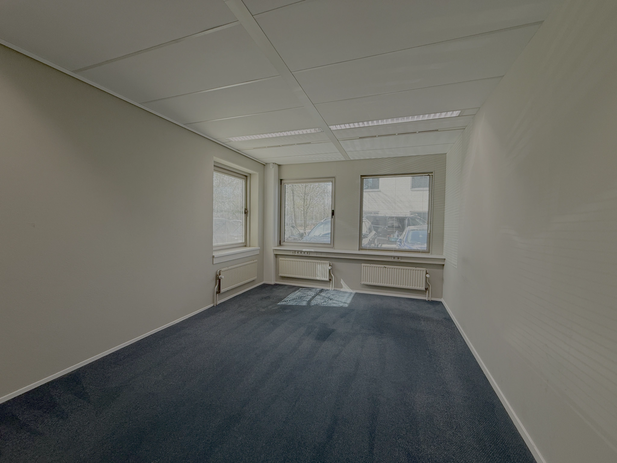 Lege kantoorruimte aan de Lunettenbaan met drie ramen, witte muren en donkerblauw tapijt.