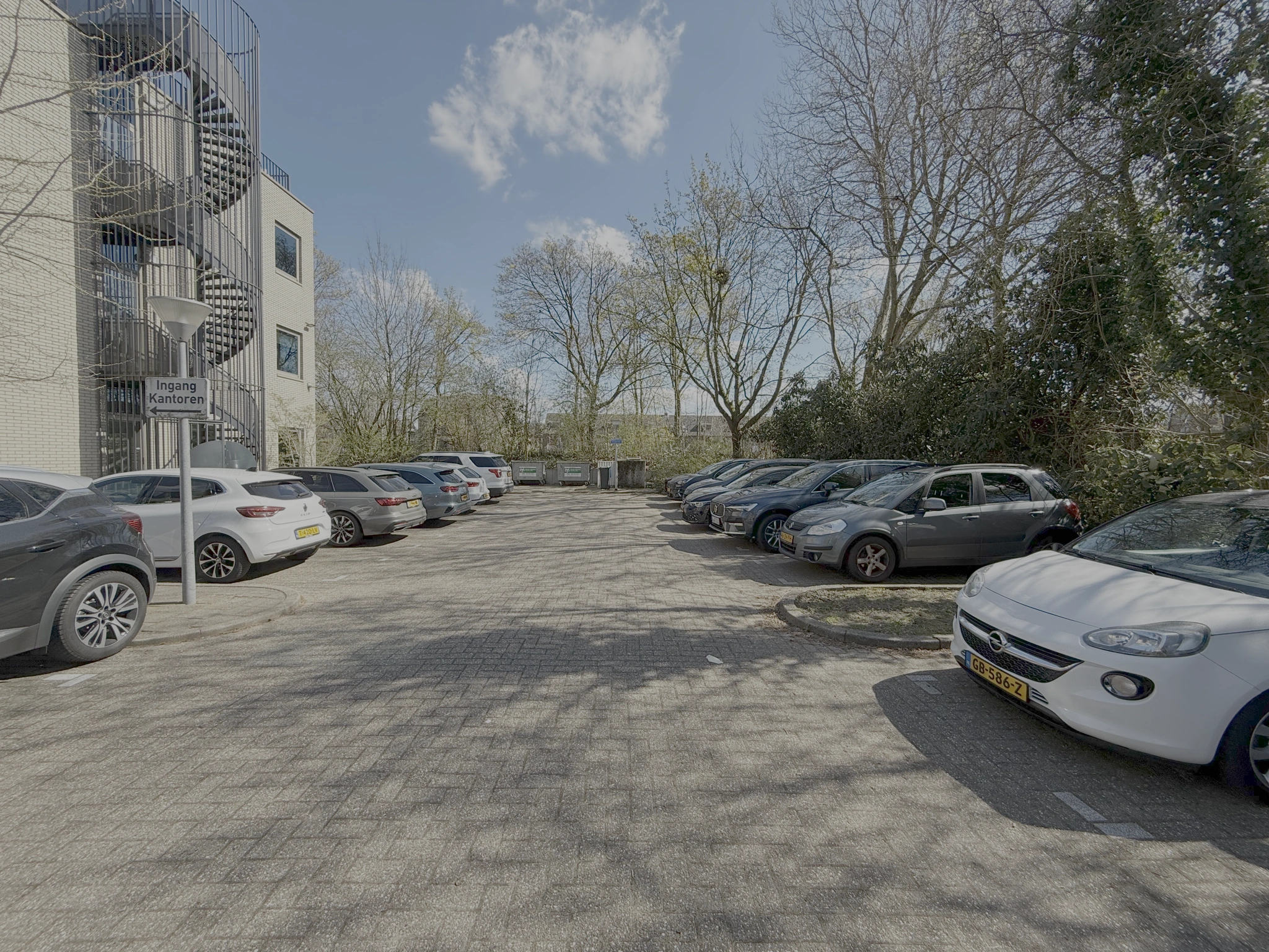 Parkeerplaats aan de Lunettenbaan met geparkeerde auto's naast een kantoorgebouw met wenteltrap en bord "Ingang Kantoren".