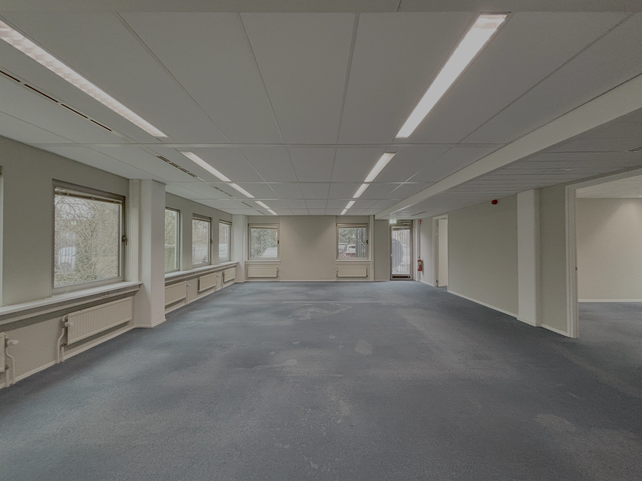 Leeg kantoorpand aan de Lunettenbaan met grote ramen, systeemplafond en blauwe vloerbedekking.