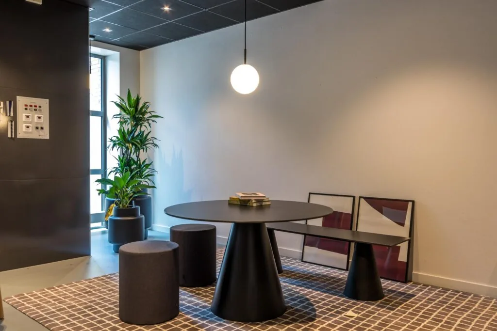 Moderne zithoek met een ronde zwarte tafel, minimalistische krukken, een hanglamp en planten in een strakke interieursetting.