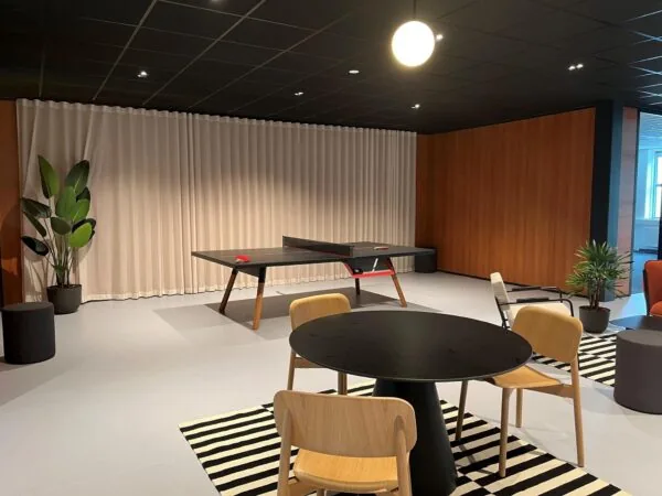 Moderne ontspanningsruimte met een pingpongtafel, ronde vergadertafel, houten stoelen en planten op een zwart-wit gestreept tapijt.