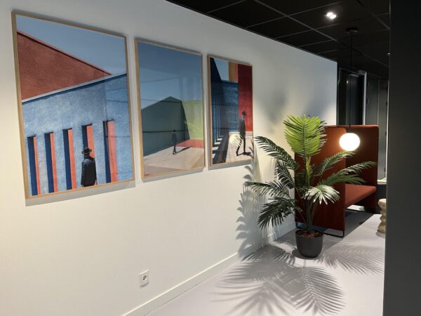 Moderne kantoorhoek met drie kleurrijke kunstwerken aan de muur, een kunstplant en een rood zitmeubel met hanglamp.