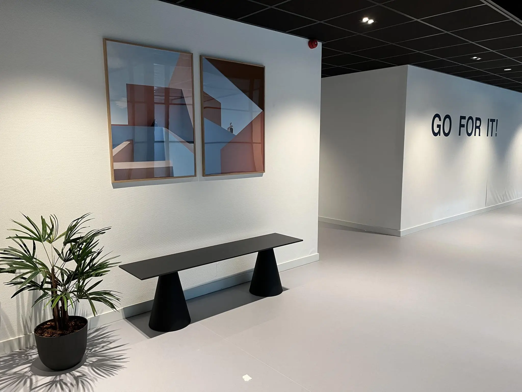 Moderne, minimalistische gang met een zwarte bank, twee abstracte kunstwerken, een kamerplant en de motiverende tekst "GO FOR IT!" op de muur.