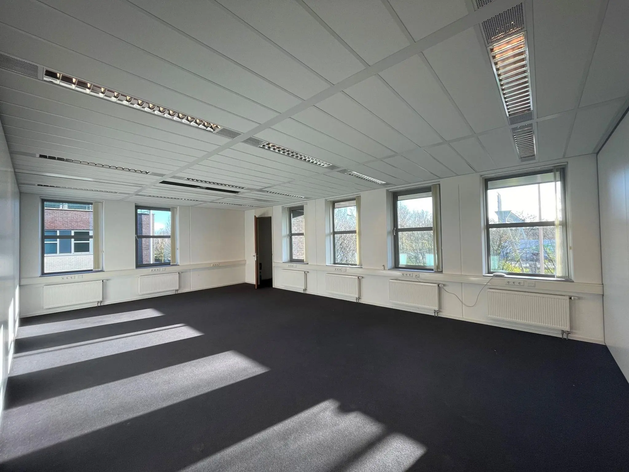 Lichte, lege kantoorruimte met vijf ramen, wit systeemplafond en donker tapijt in gebouw De Wel.